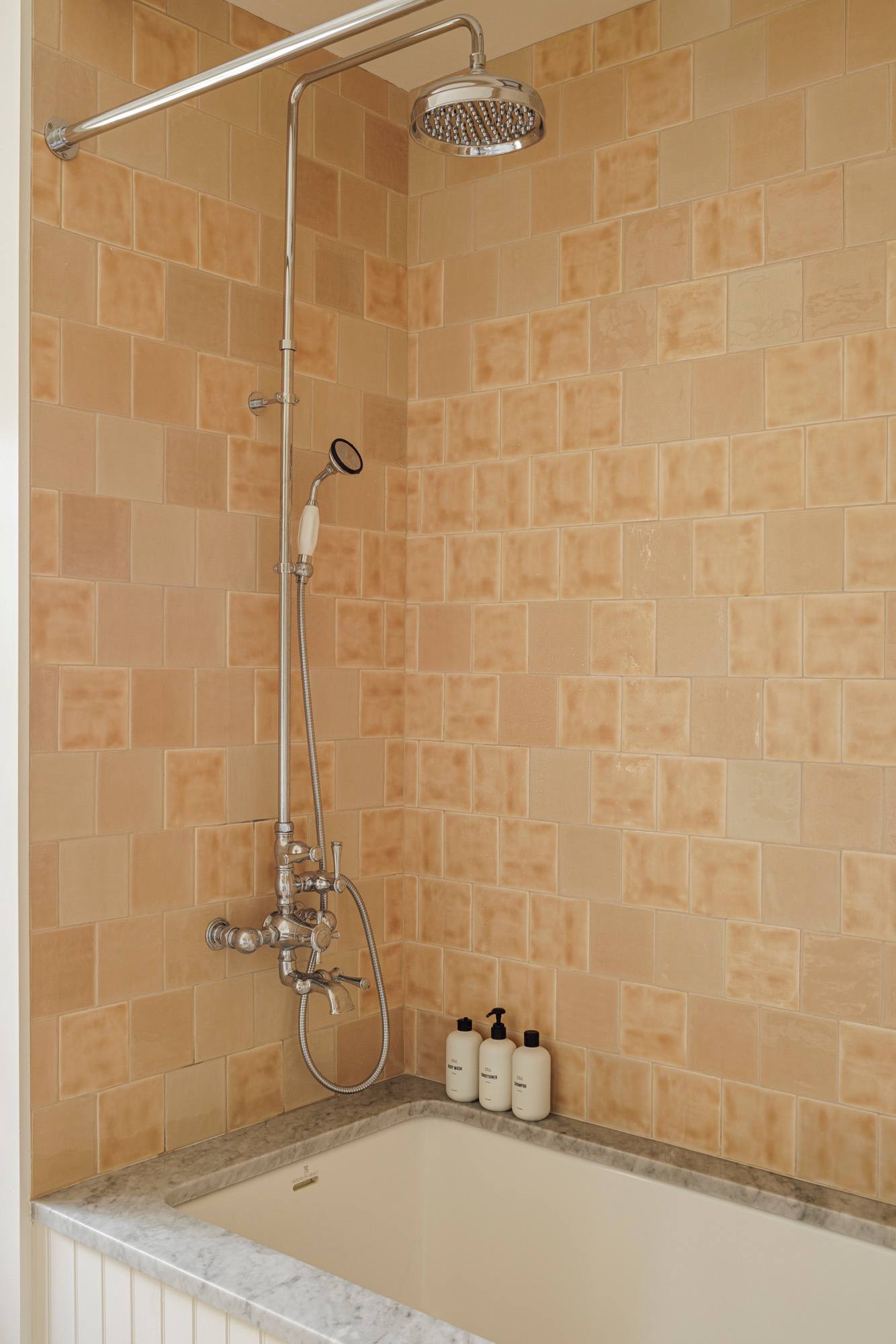 A bathroom featuring a bathtub with light orange square tiles on the wall and a chrome shower system, including a rain shower head and a handheld sprayer. A marble ledge holds bath products.