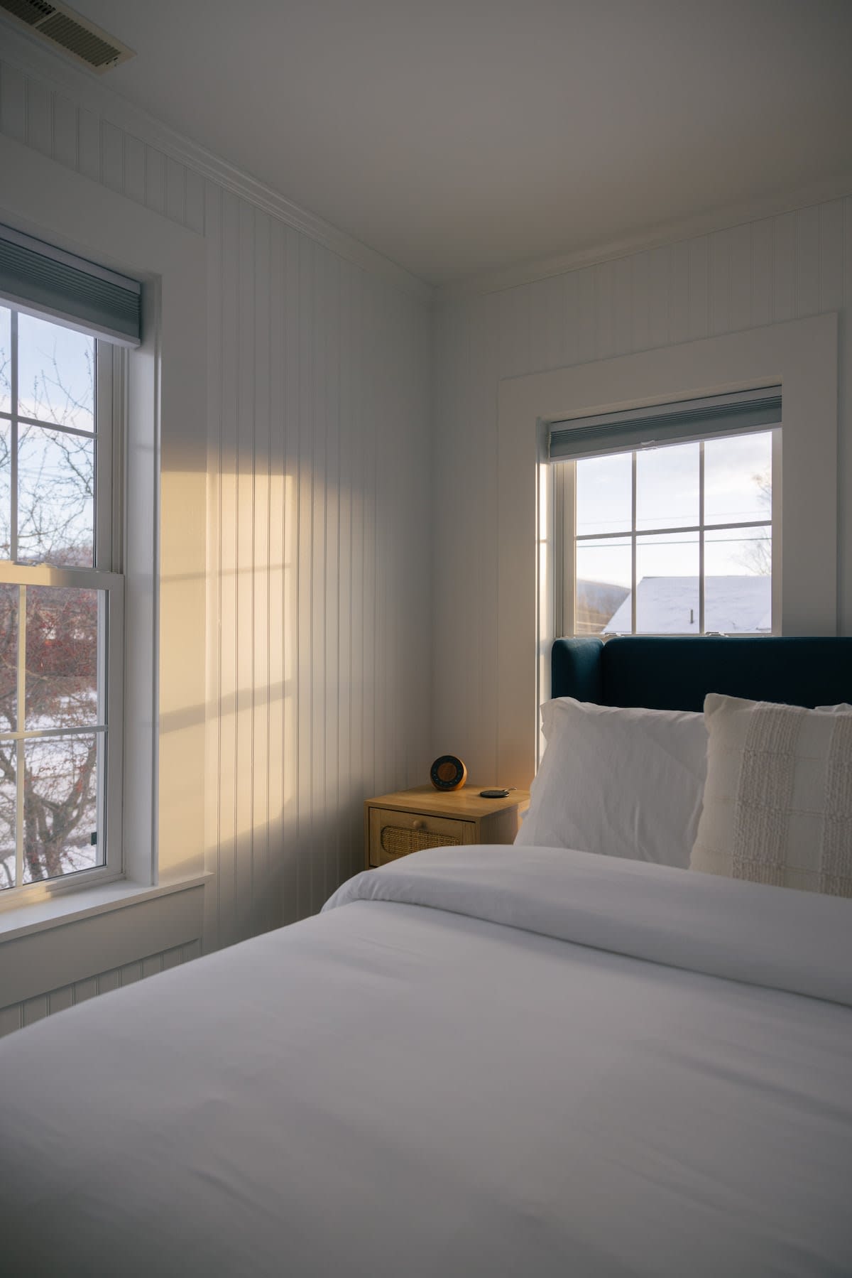 Sunlit bedroom with white vertical paneling walls, a bed with a blue upholstered headboard, white bedding, and a wooden nightstand beside a window with warm light streaming in.