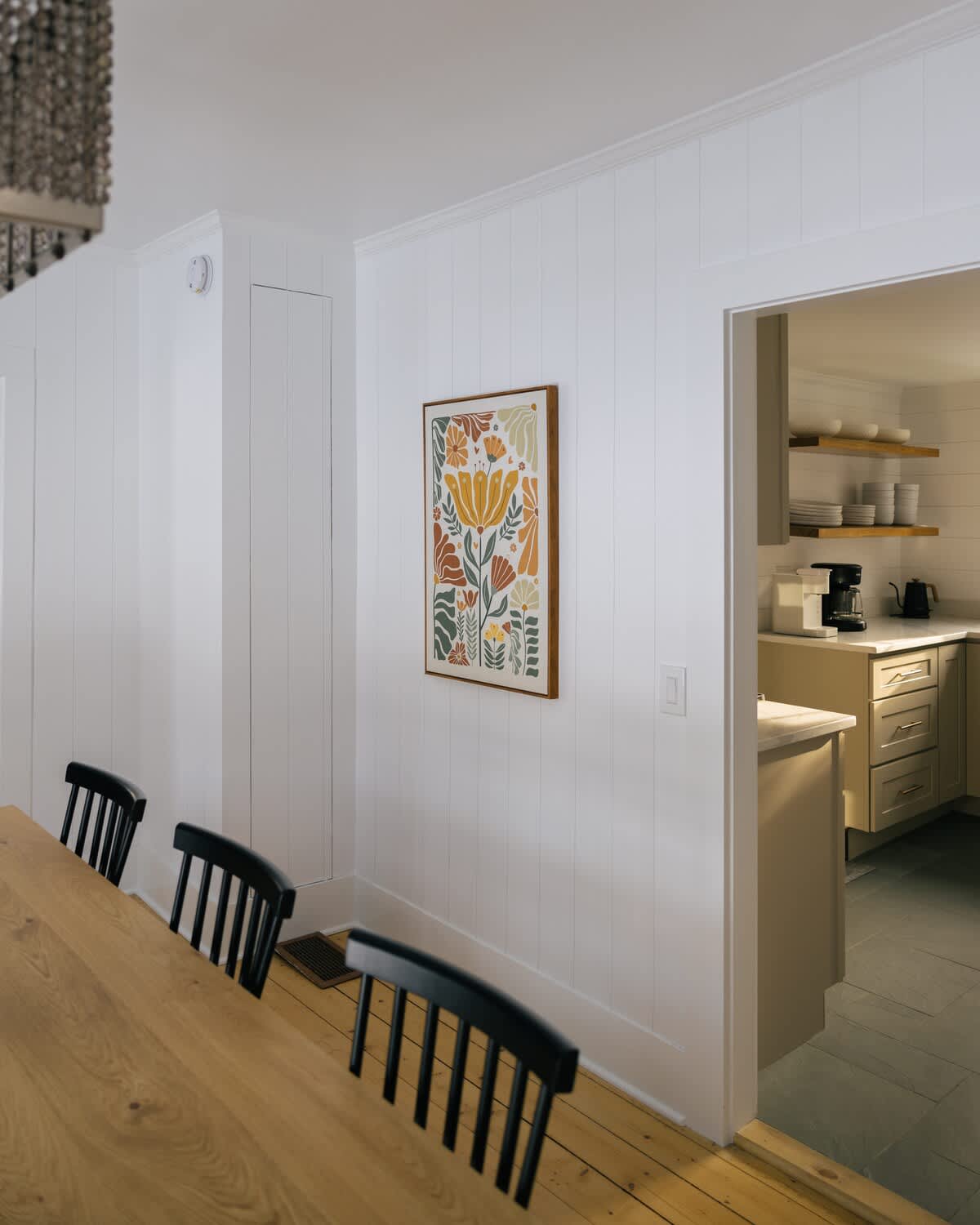 A dining room wall adorned with a colorful floral art print, white shiplap paneling, and a doorway leading into the kitchen, revealing open shelves and appliances.