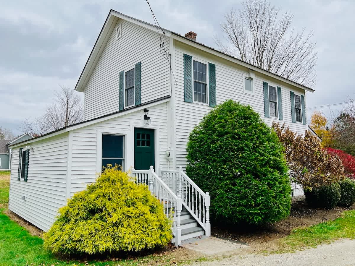 White farmhouse exterior with dark green shutters and a green front door, featuring a small porch with white railings and vibrant green and yellow shrubs in the foreground.