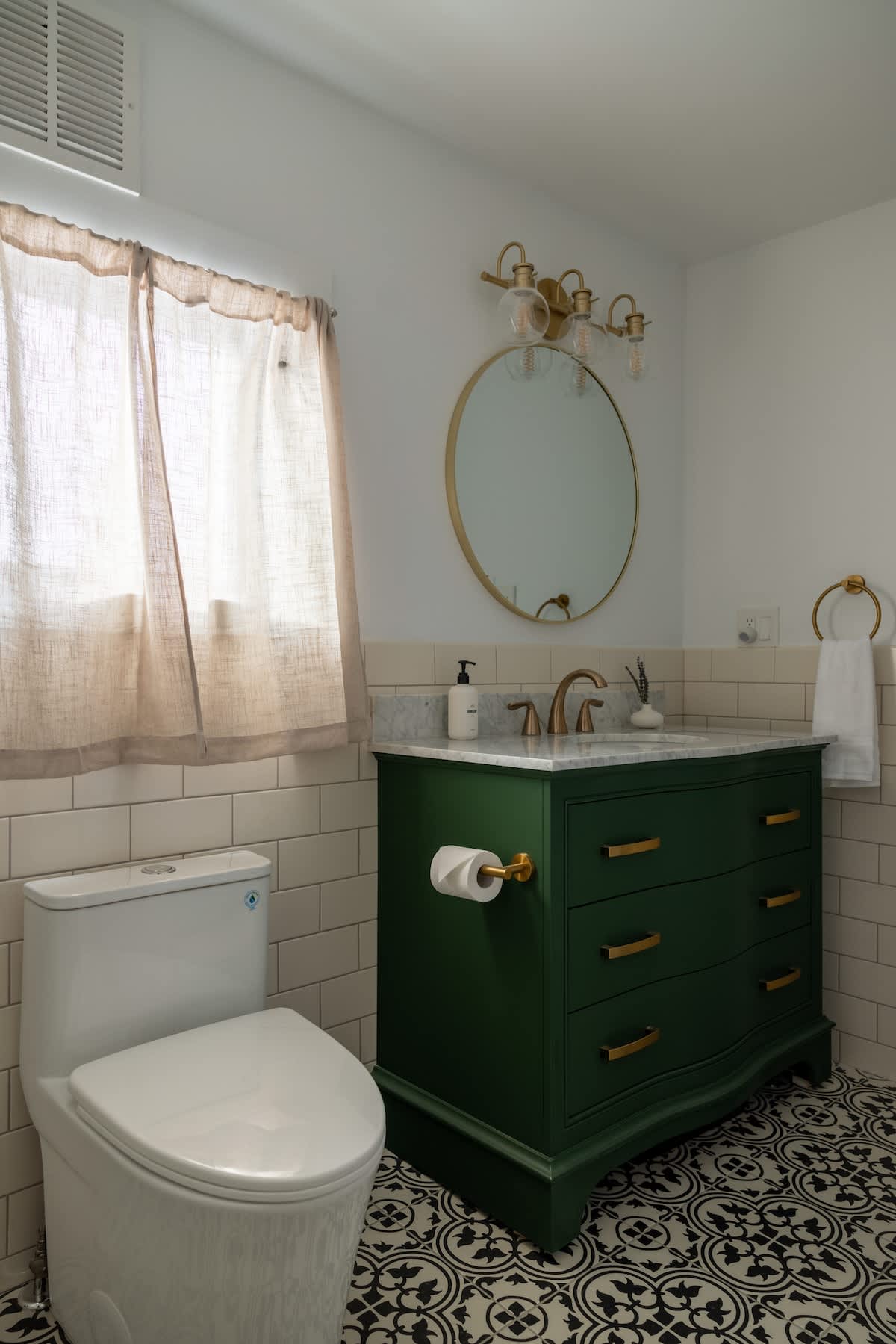 Modern bathroom featuring a dark green vanity with a marble top and gold hardware, a round gold-framed mirror, subway tiled walls, and a striking black and white patterned floor.