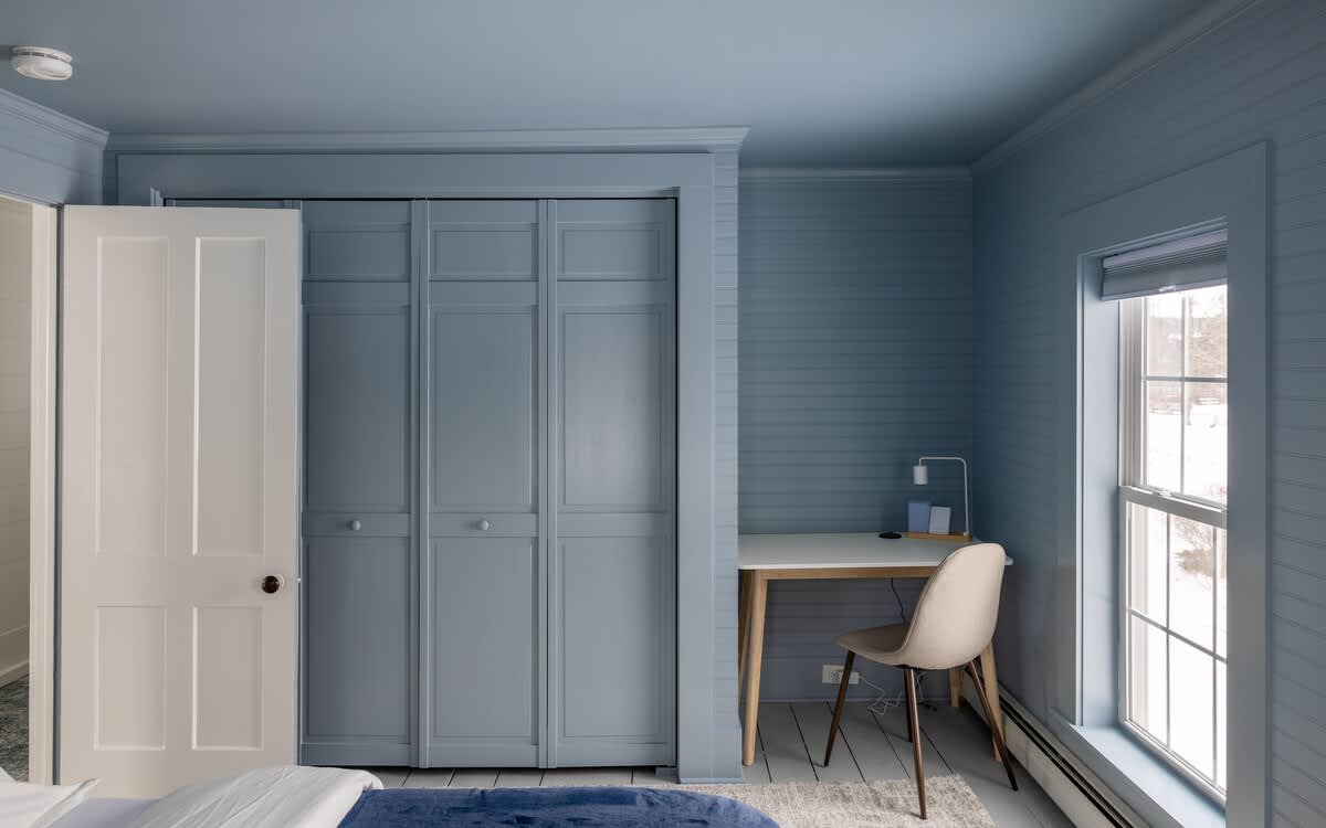 Another view of the blue-painted bedroom, showing a small desk with a chair next to a window, and a blue bi-fold closet door, maintaining the consistent shiplap wall texture.