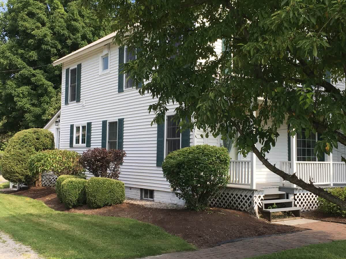 Charming white farmhouse exterior with dark green shutters, a covered porch with white railings, and lush green landscaping with mature trees and shrubs under a sunny sky.