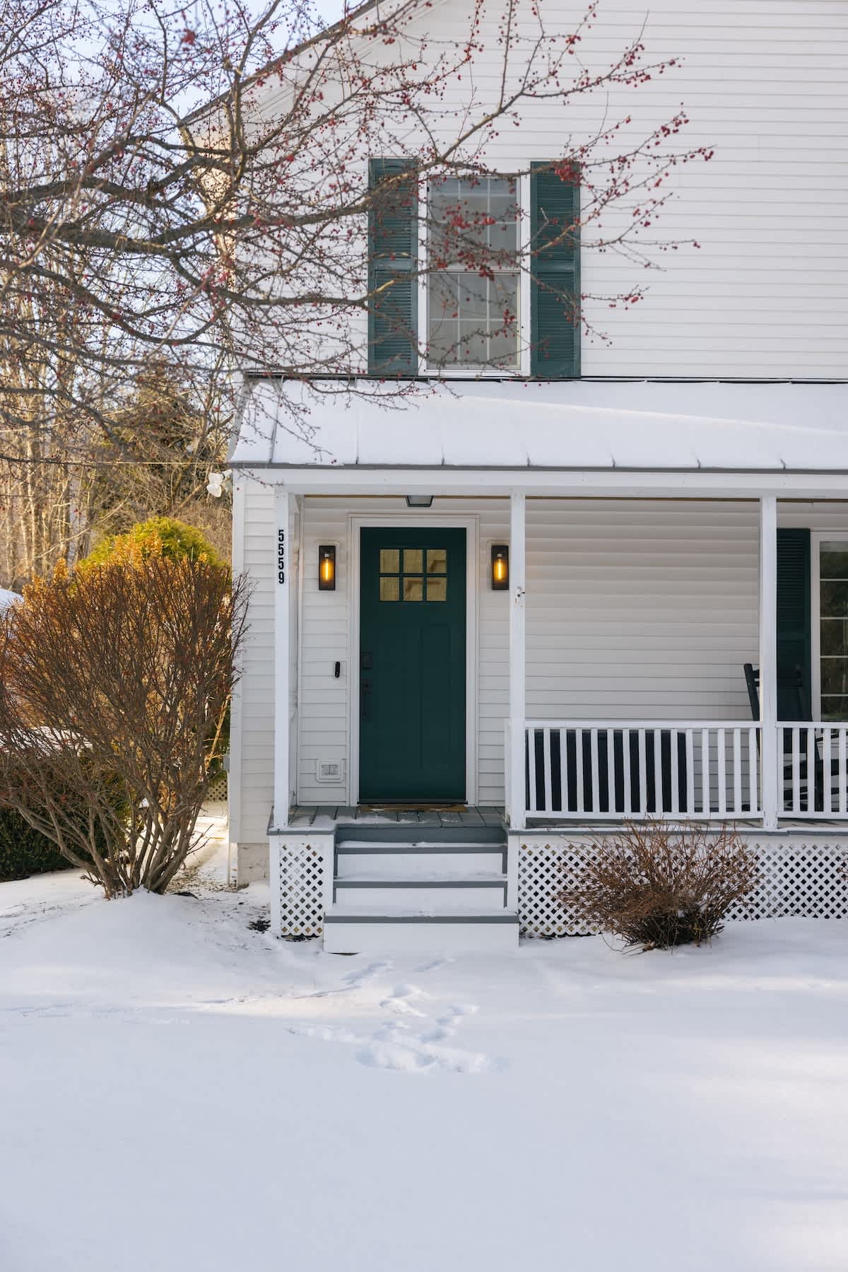 An inviting front porch features a dark green door flanked by two modern sconce lights, white railings, and steps leading to a snow-dusted path with visible footprints.