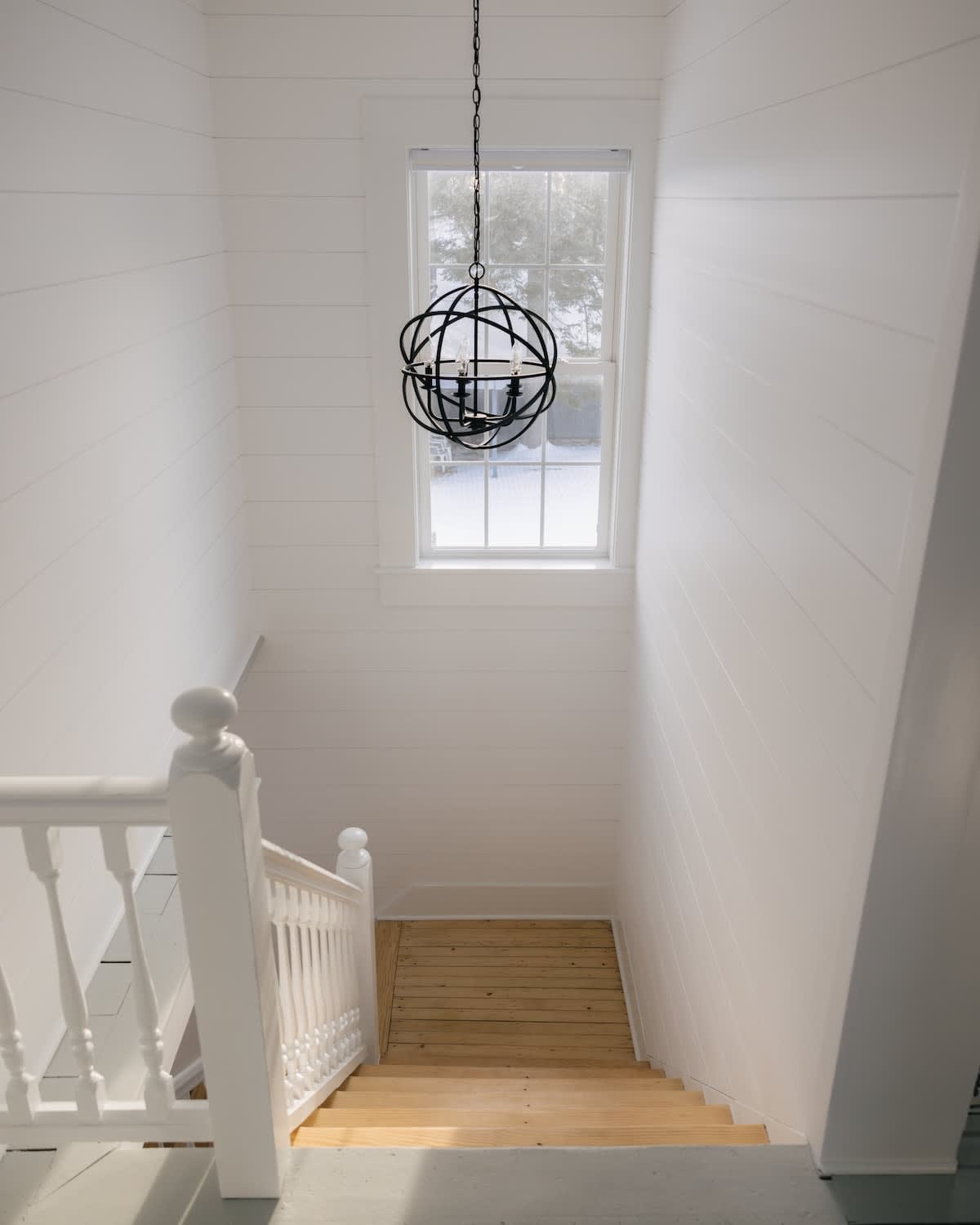 Bright staircase area with white horizontal shiplap walls, white banisters, light wood stairs, and a striking black spherical chandelier hanging in front of a window.