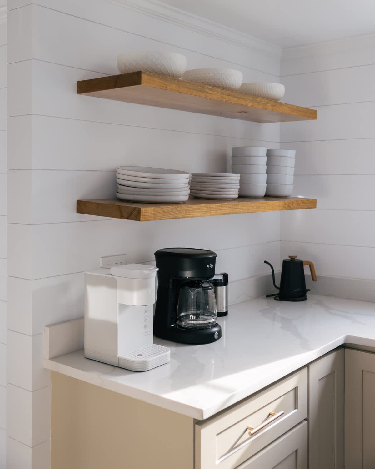 A bright kitchen corner with white shiplap walls, light wood open shelving displaying white dishes, and a white countertop holding a white single-serve coffee maker, a black drip coffee maker, and a black kettle.