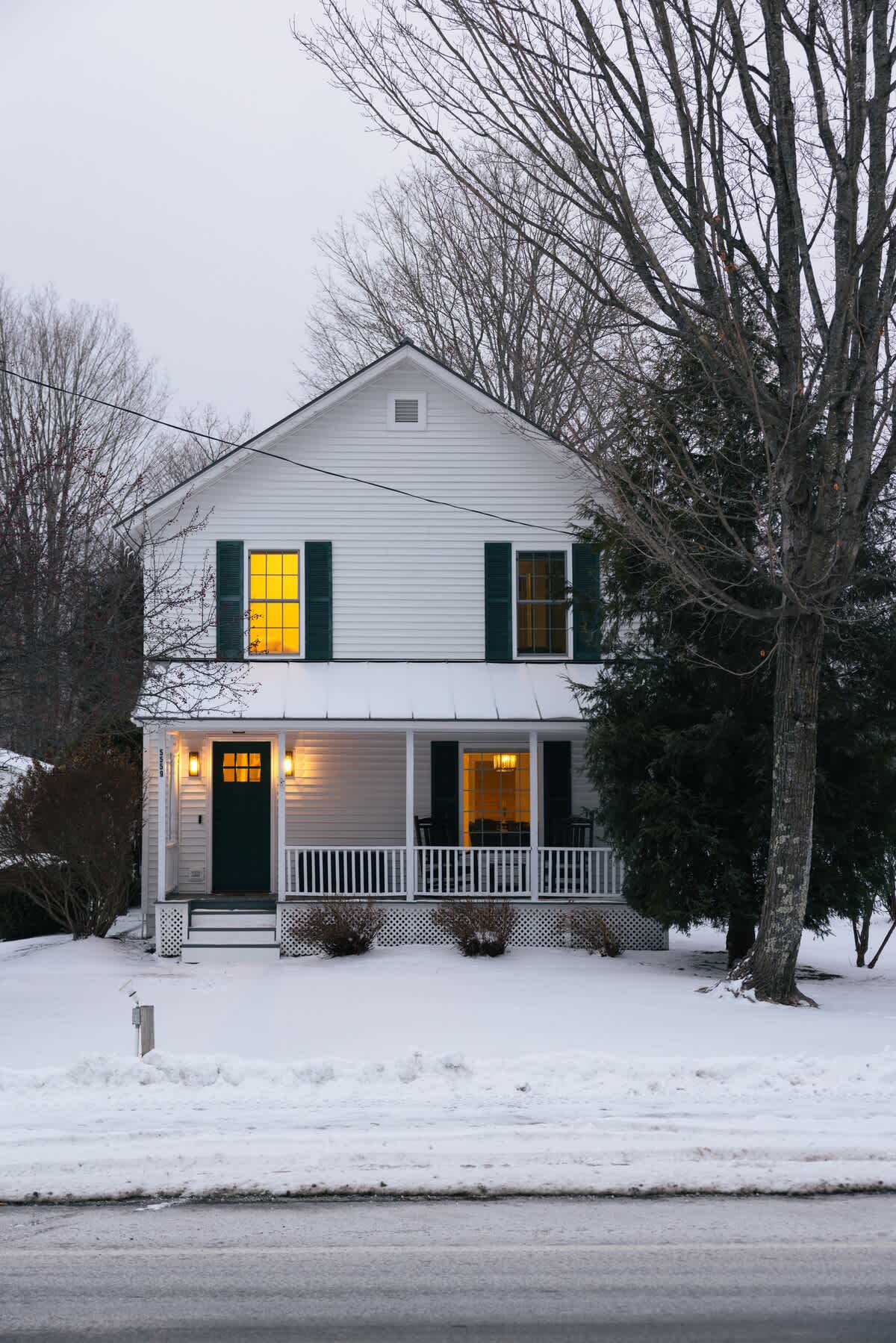 Picturesque white farmhouse exterior covered in snow at dusk, with warm lights glowing from inside the windows and a welcoming light by the green front door, framed by bare trees.