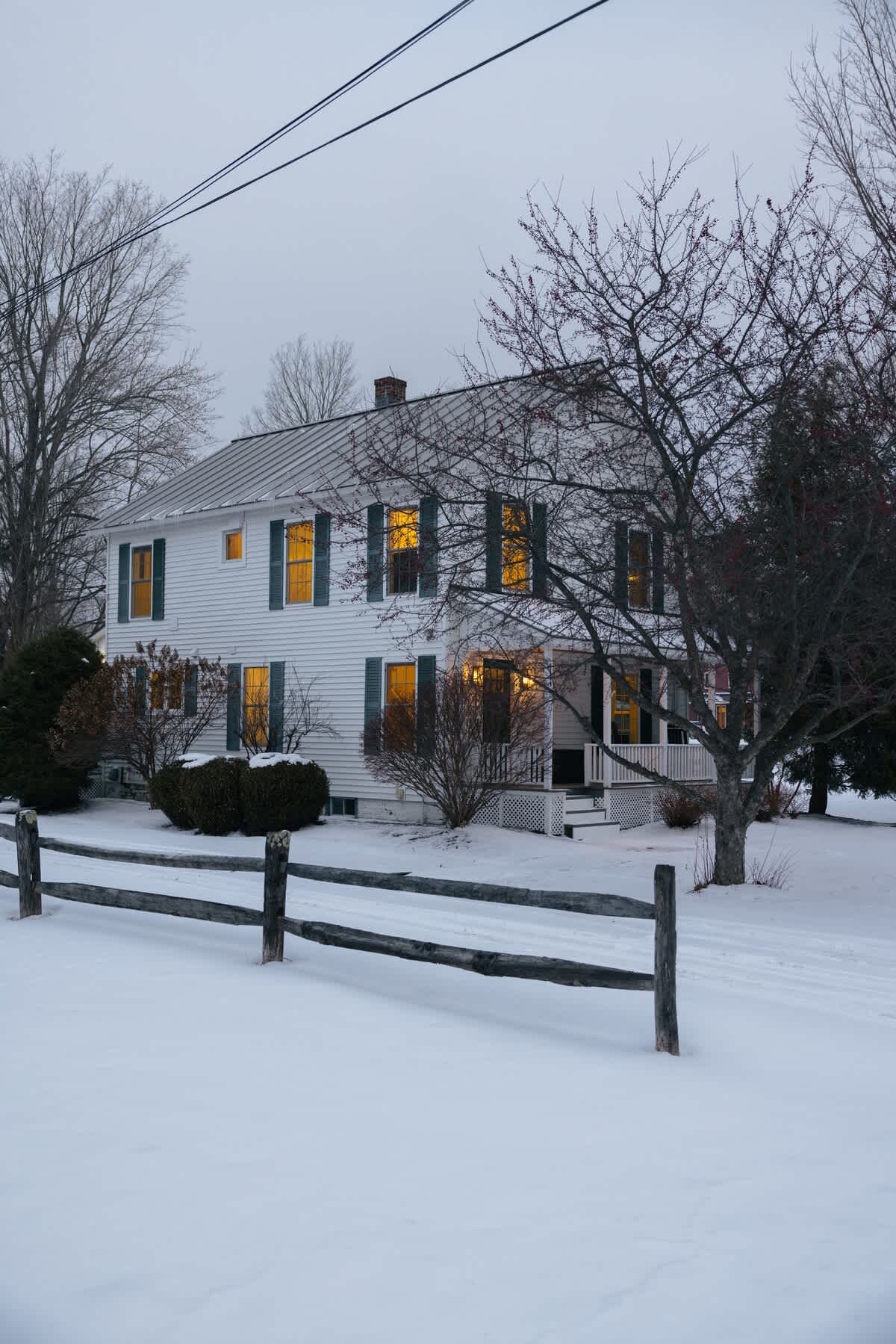 A charming white farmhouse with dark green shutters and a metal roof, nestled in a snow-covered yard with a rustic split-rail fence and mature trees, illuminated by warm interior lights.