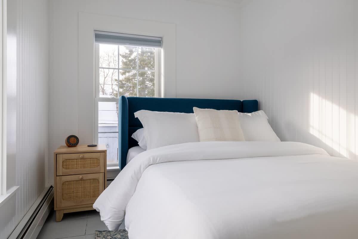 Cozy bedroom featuring white vertical paneling walls, a bed with a blue upholstered headboard, white bedding, and a wooden nightstand with woven drawers next to a window.