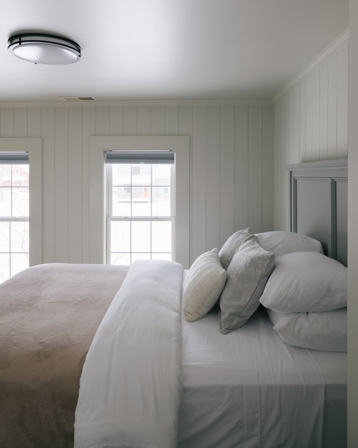 Bright bedroom with white vertical paneling walls, a bed with white and beige bedding, and two large windows offering natural light. A modern flush-mount light fixture is on the ceiling.