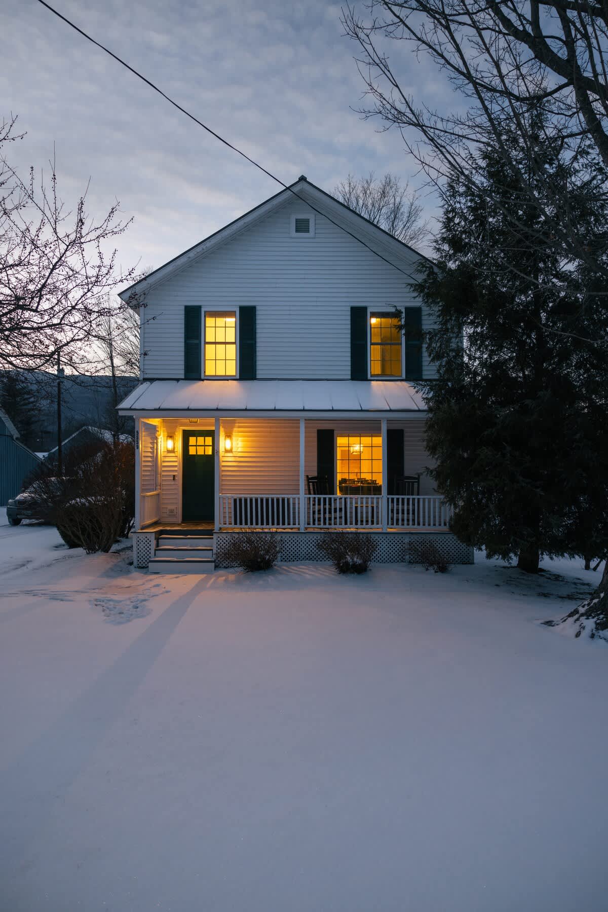 A classic white clapboard farmhouse with dark green shutters and a welcoming front porch, illuminated by warm interior lights, stands in a snow-covered yard at dusk with bare trees framing the scene.