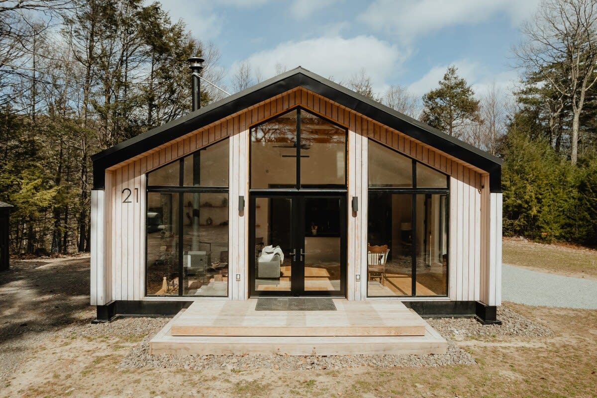 The modern front facade of a cabin features a striking A-frame design with extensive floor-to-ceiling windows and a central double door, set against light wood siding and a black roof, surrounded by a natural, wooded landscape.