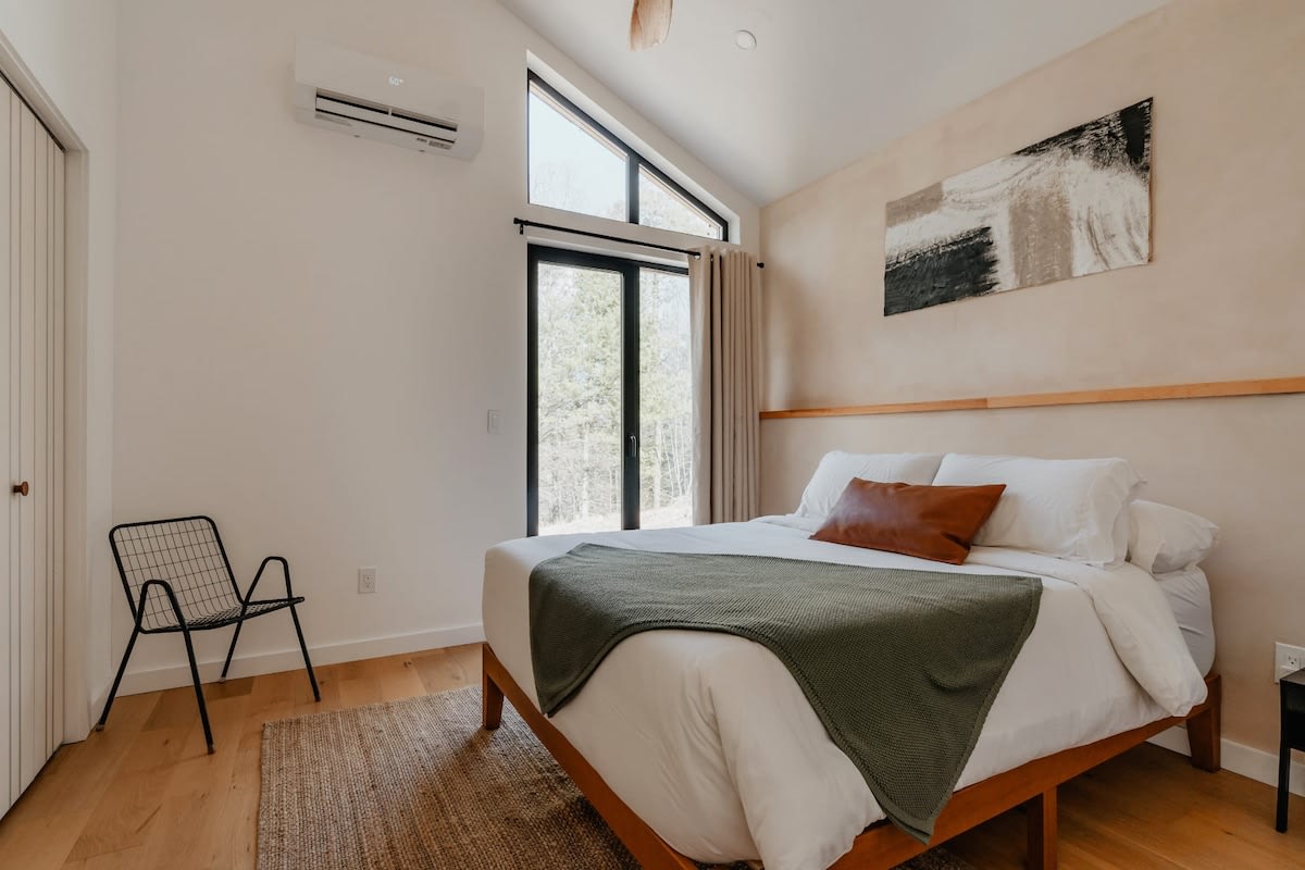 Bright and airy bedroom featuring a queen-sized bed with white linens, a green throw, and a brown accent pillow. Large angled windows offer views of the outdoors, complemented by light wood flooring and a modern black wire chair.