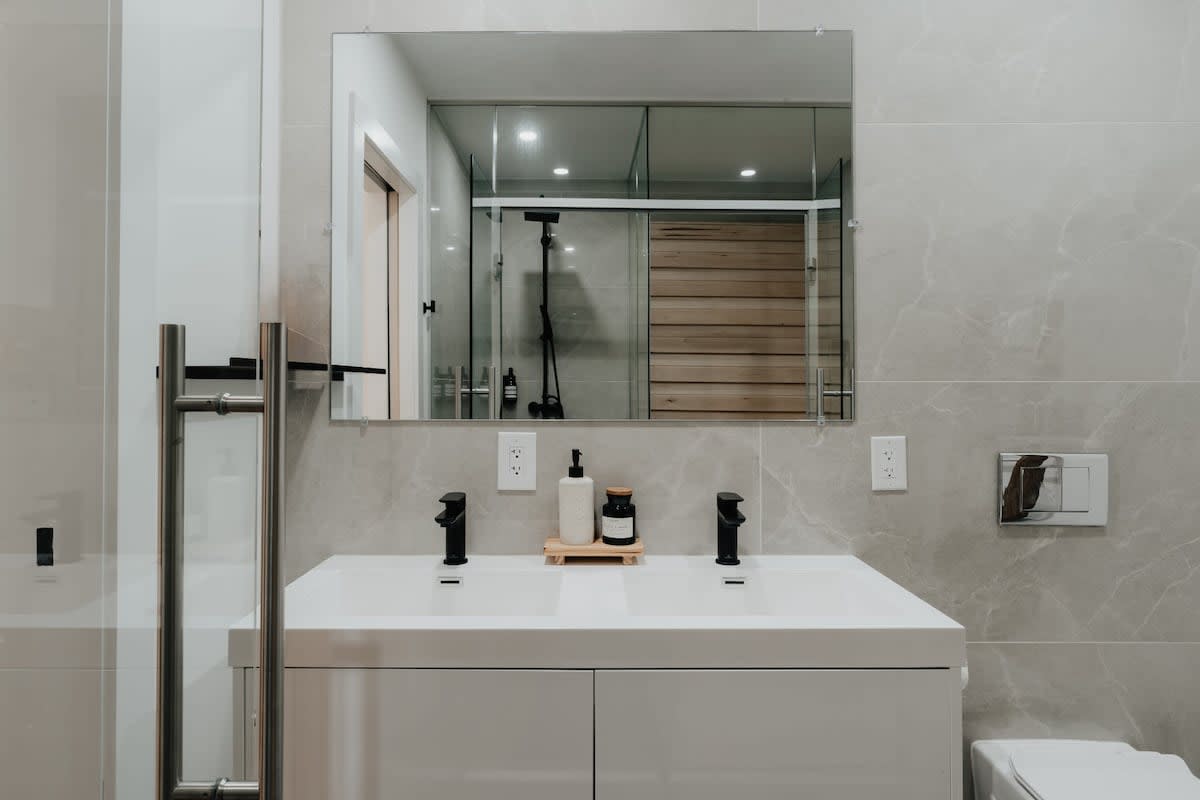 Spacious bathroom with a wide mirror reflecting the wooden sauna and glass shower enclosure. The room includes a white double vanity with black faucets and light gray tiled walls, creating a clean and modern aesthetic.