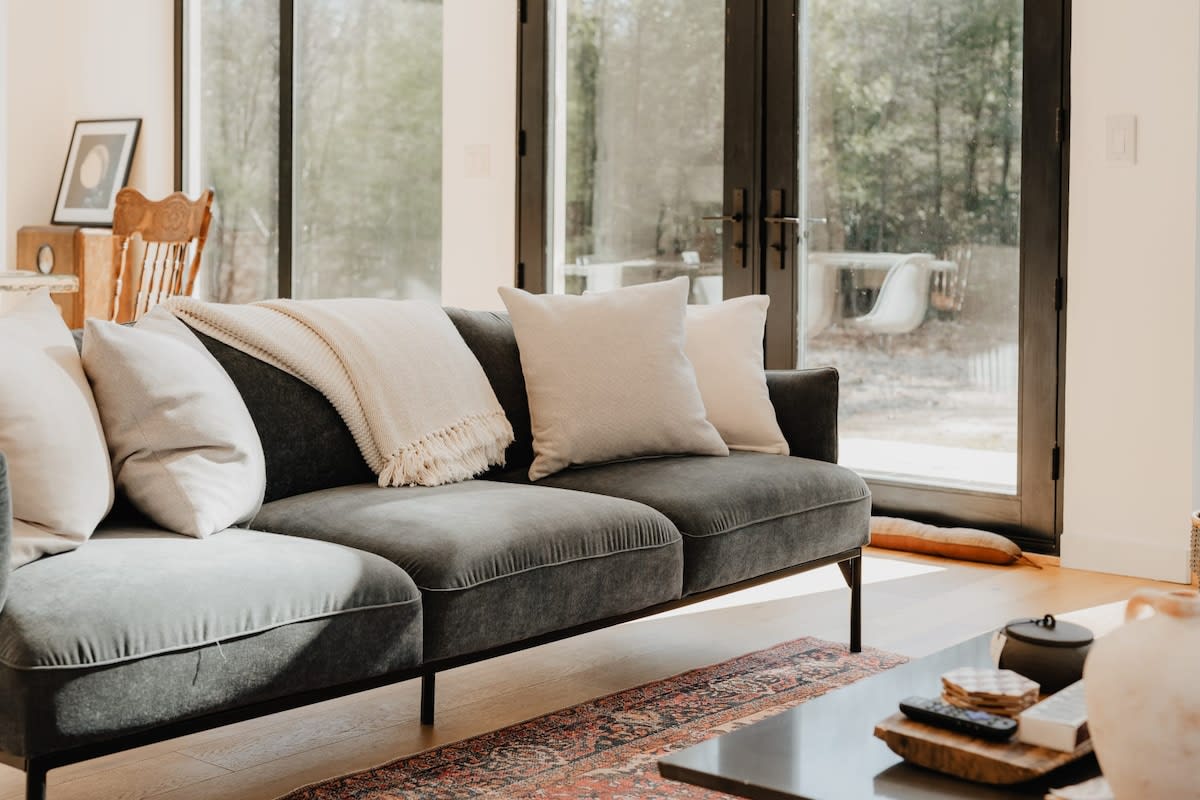 A comfortable grey velvet sofa with light-colored throw pillows and a blanket, positioned on a patterned area rug. Large black-framed glass doors and windows offer a view of the outdoor area and surrounding trees.