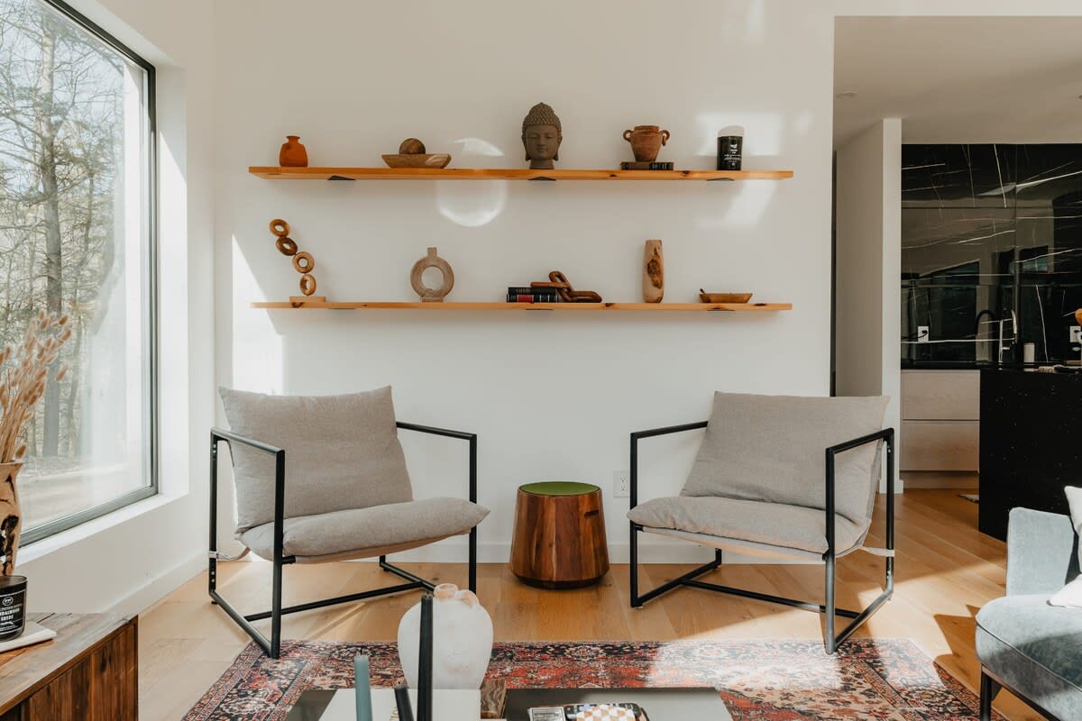 A bright living room corner with two modern armchairs featuring light grey cushions and black frames, separated by a round wooden side table. Two floating wooden shelves on the white wall display various decorative objects, including a Buddha head.