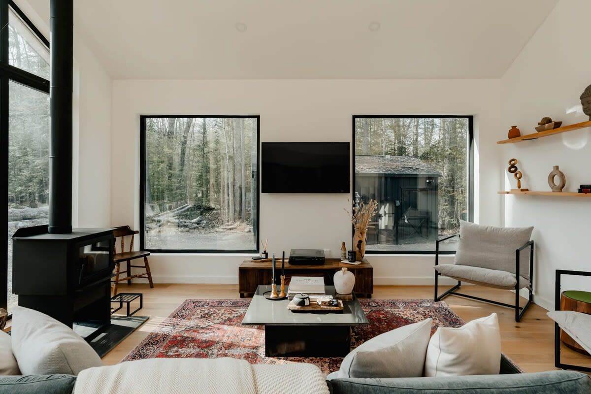 A modern living room with white walls, light hardwood floors, and a large patterned rug. It features a black wood-burning stove, a wall-mounted TV, and large windows offering views of a wooded area.