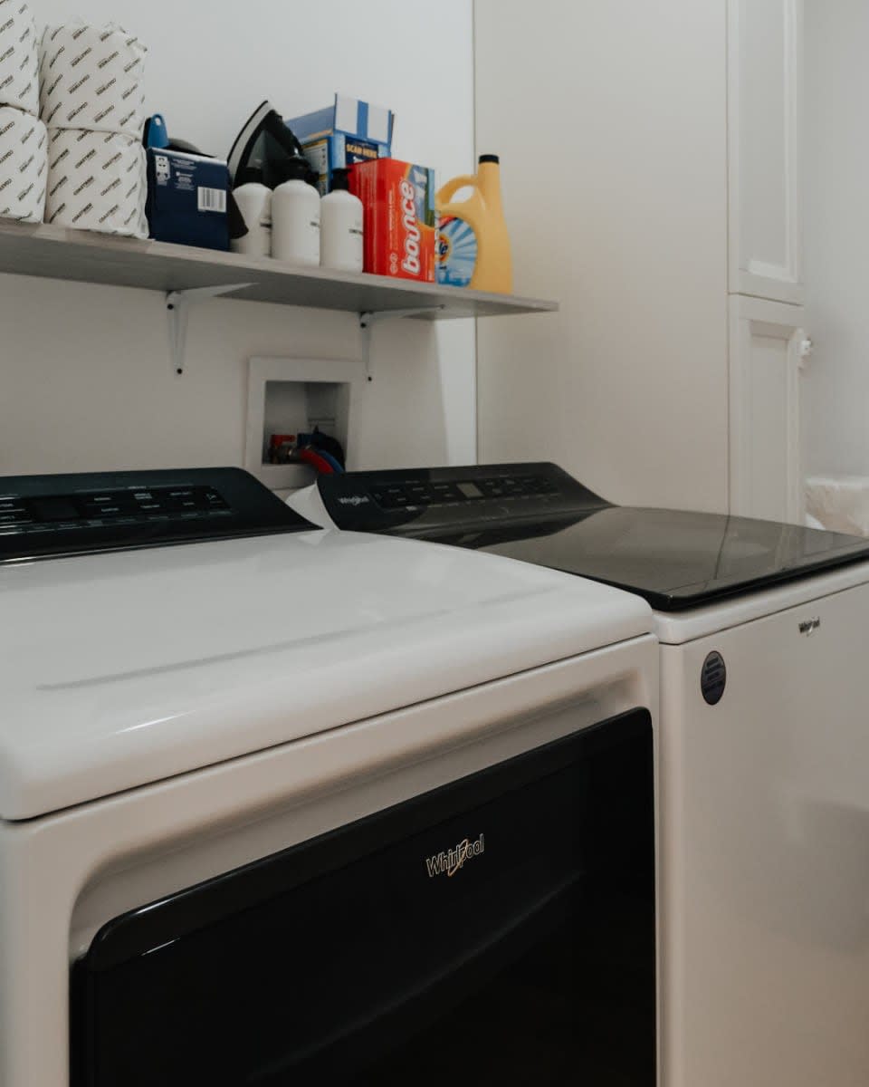 A functional laundry room with a white top-loading washing machine and a black-front dryer, topped with a grey shelf holding various laundry supplies like detergent, dryer sheets, and toilet paper.