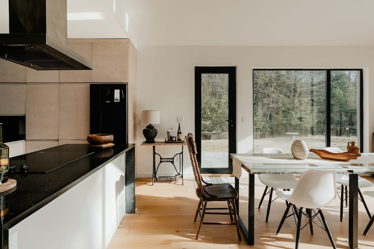 A modern kitchen area with sleek black countertops and light wood cabinetry, featuring a black refrigerator and a range hood. A rustic side table with a lamp stands next to a black-framed glass door, leading to the outdoors.