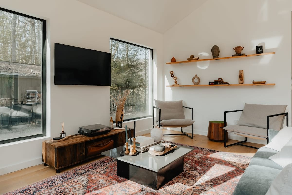A bright living room with light hardwood floors and a large patterned rug, featuring a wall-mounted TV above a wooden console. Two modern armchairs flank a wooden side table, with large windows showcasing the surrounding forest.
