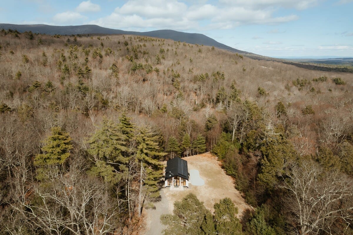 A high aerial view captures the modern cabin nestled within a vast, rolling wooded landscape, surrounded by a mix of evergreen and deciduous trees, with distant mountains visible under a partly cloudy sky.