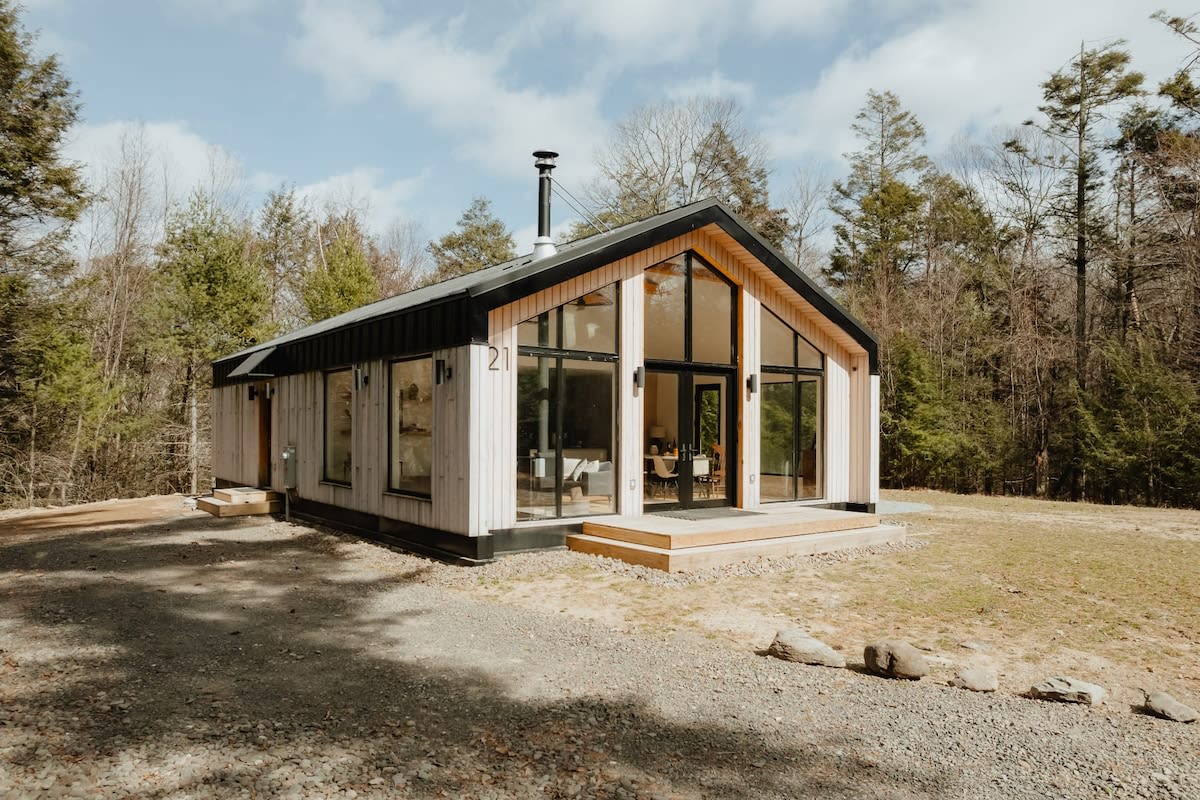 The front exterior of the modern cabin features light wood siding and a dark roof, with prominent large windows and glass doors, surrounded by a sparse forest in early spring. A gravel driveway leads up to the house.