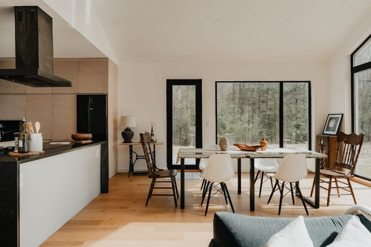An open-concept view showing the dining area with a rustic table and mixed chairs, adjacent to a modern kitchen with black countertops and light wood cabinets. Large windows and a glass door connect the space to the natural surroundings.
