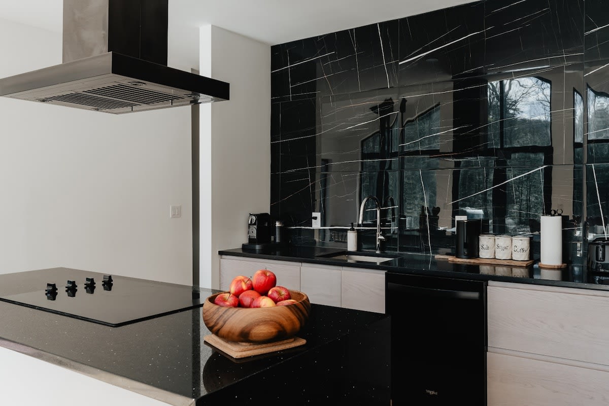 Contemporary kitchen with a large island featuring a built-in stovetop and black speckled countertop, adorned with a wooden bowl of red apples. The space boasts a dramatic black marble backsplash and light wood cabinetry, with a stainless steel range hood overhead.