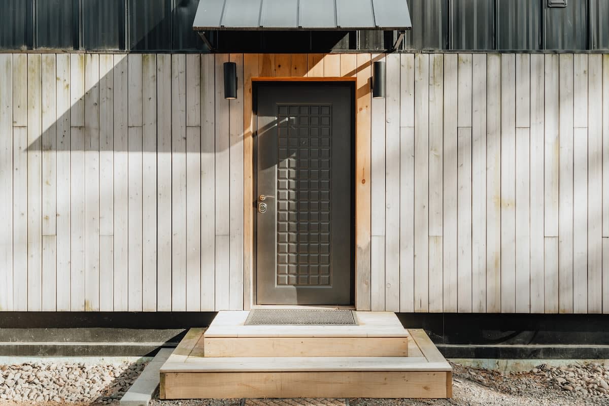 The side entrance of the cabin showcases a dark grey door with a distinctive grid pattern, illuminated by black wall sconces, set against light wood siding. A small wooden step provides access to the entrance.
