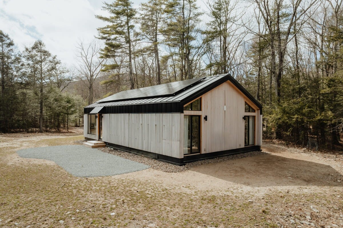 An aerial perspective reveals the modern cabin's elongated structure with light wood siding, a black roof, and solar panels, situated amidst a sparse, wooded landscape with a gravel area beside it.
