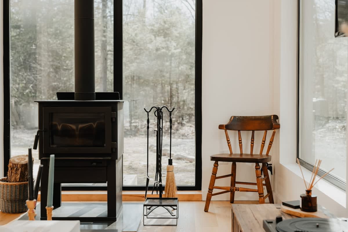 A close-up of the black wood-burning stove with a tall black flue, set on light hardwood floors. A rustic wooden chair and a set of black fireplace tools are positioned nearby, with large windows revealing a snowy wooded landscape.