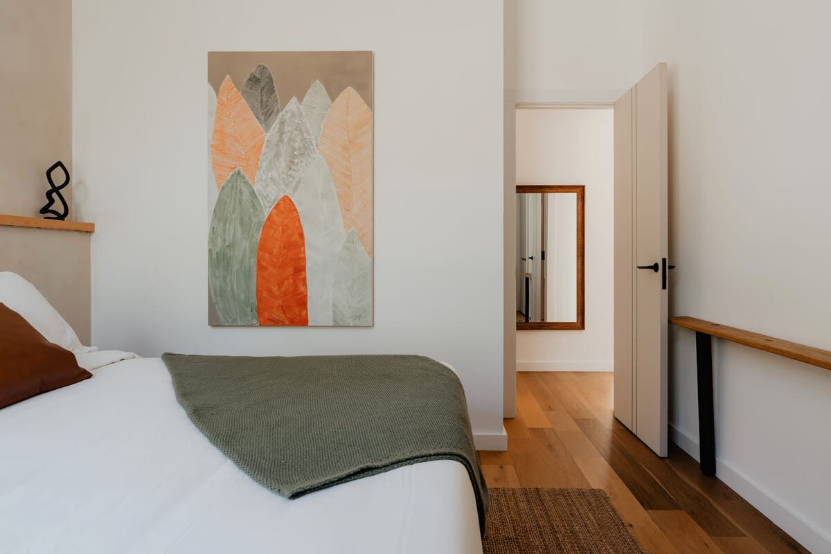 View of the bedroom showing a bed with a green throw and a vibrant abstract painting featuring orange, green, and gray leaf-like shapes. The room has light wood flooring and a doorway leading to another area, with a full-length mirror visible beyond.