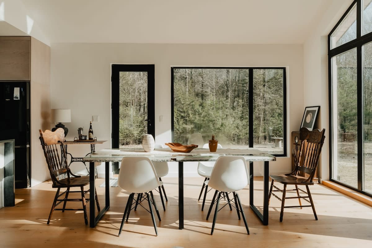 A bright dining area featuring a rustic wooden table with black metal legs, surrounded by a mix of modern white chairs and traditional wooden chairs. Large black-framed windows and a glass door provide expansive views of the surrounding forest.