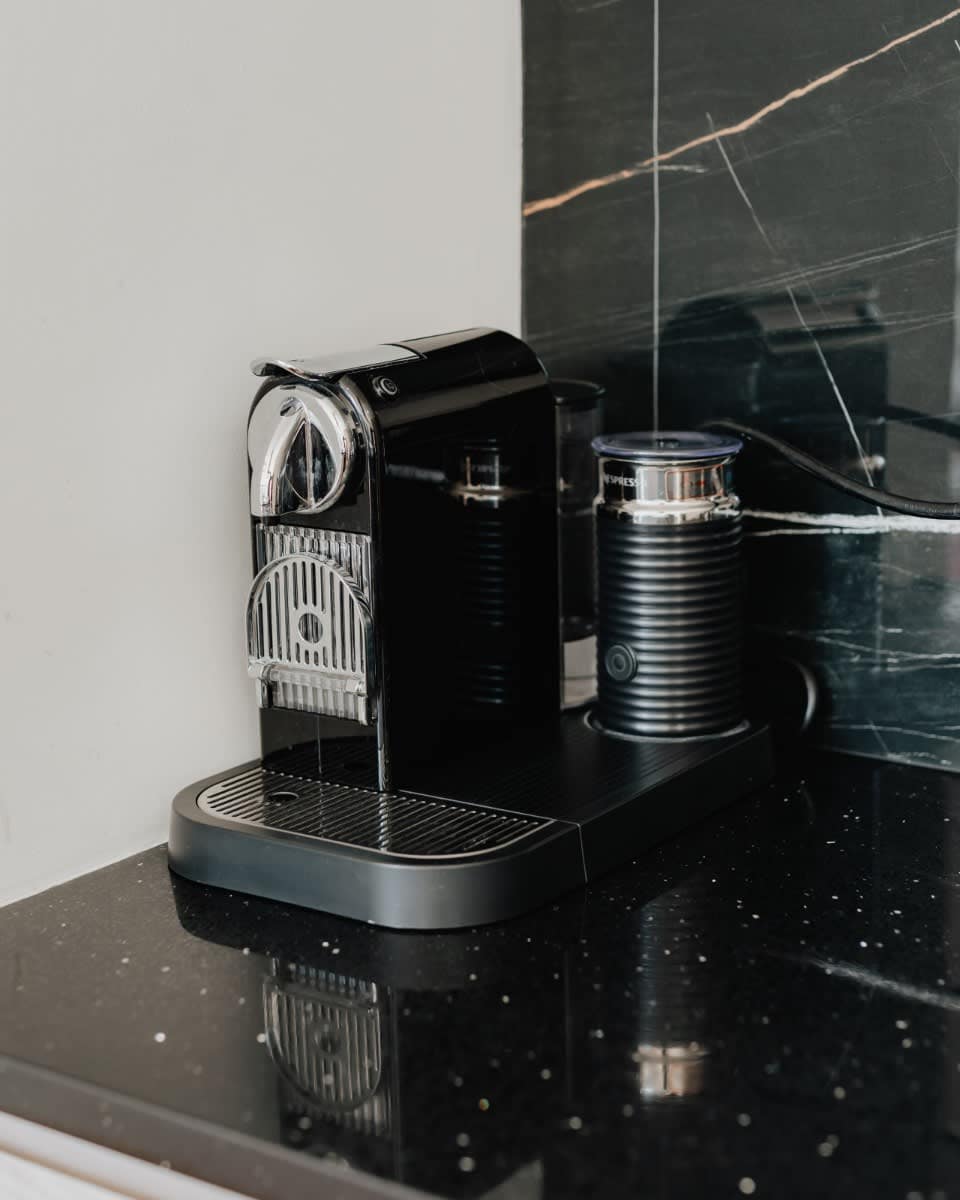 A sleek black Nespresso coffee machine and milk frother sit on a black speckled countertop, set against a dramatic black marble backsplash. The modern appliances are ready for use, adding a touch of luxury to the kitchen.
