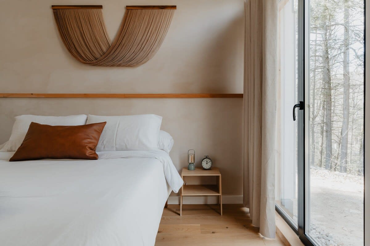 A modern bedroom with a white bed, brown leather pillow, and green throw, featuring a light-colored wall with a wooden shelf and a large woven art piece. A small wooden nightstand with an alarm clock and lamp sits beside the bed, next to a large window with beige curtains overlooking a wooded area.