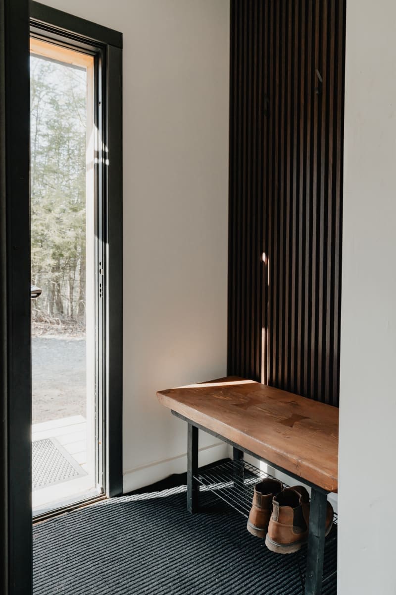A bright entryway features a wooden bench with a metal frame, a black mat, and a pair of boots, set against a white wall with a dark wood slatted accent wall. A glass door opens to an outdoor, wooded setting.
