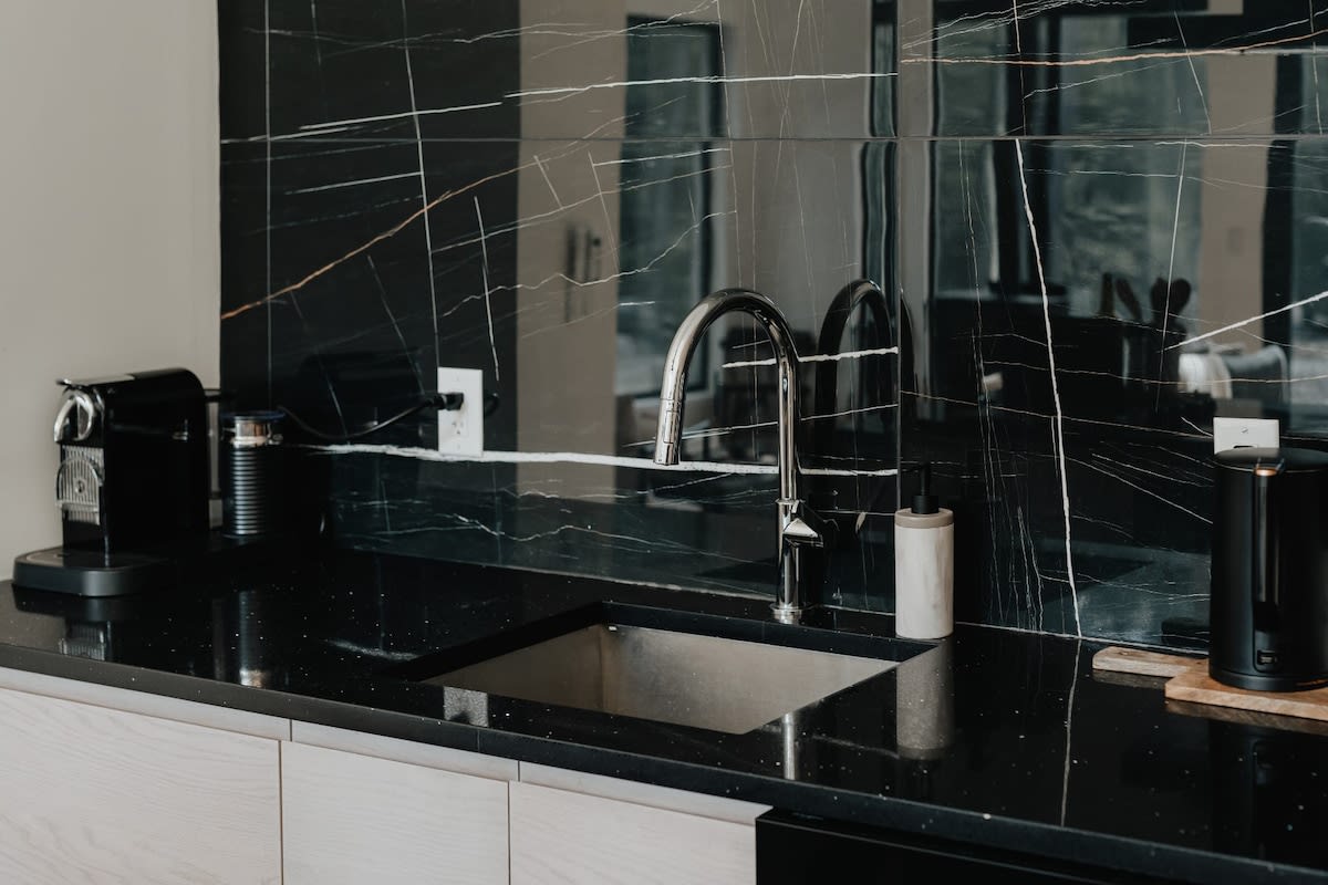 Close-up of the kitchen sink area, showcasing a deep black speckled countertop and a square stainless steel sink with a modern chrome gooseneck faucet. The bold black marble backsplash reflects the room, and a black coffee machine sits nearby.