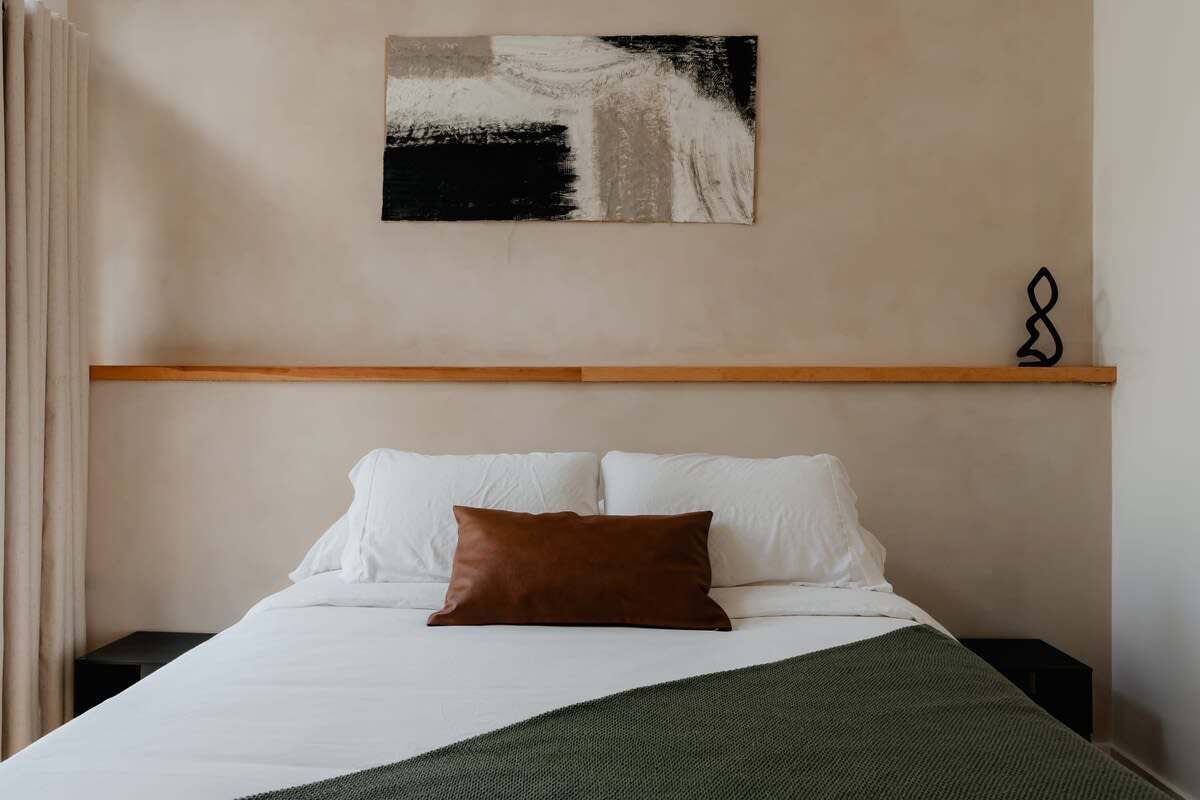 A cozy queen bed with crisp white bedding, a rich brown leather accent pillow, and a textured green throw blanket. Above the bed, an abstract black and white painting hangs over a minimalist wooden shelf, adding a touch of modern art.