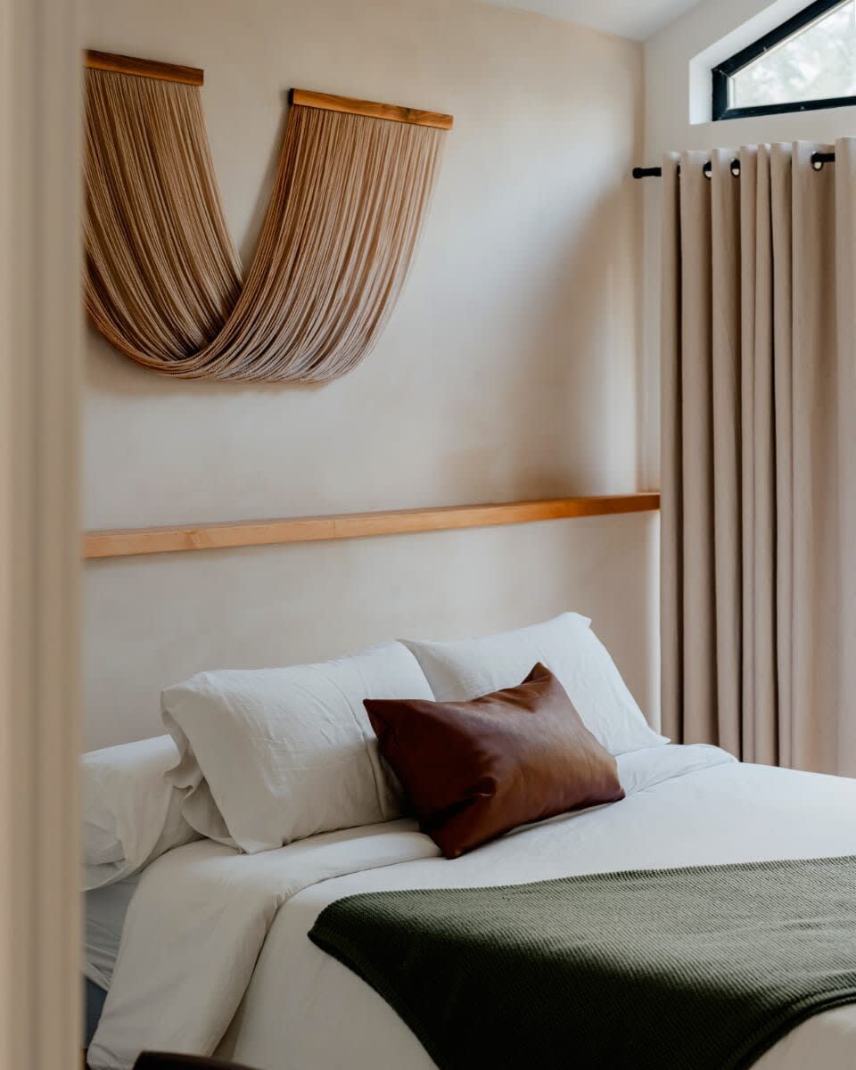 A cozy bedroom features a white bed with a brown leather pillow and a green throw blanket, set against a light-colored wall with a wooden shelf and a unique woven wall hanging. A small window is visible in the upper right.