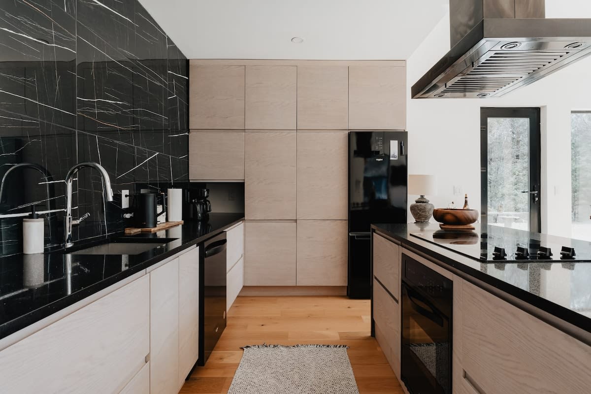 Modern kitchen featuring a striking black marble backsplash with white veining, black speckled countertops, and light wood flat-panel cabinetry. A black refrigerator and sleek black appliances complement the dark surfaces, while a light wood floor adds warmth.