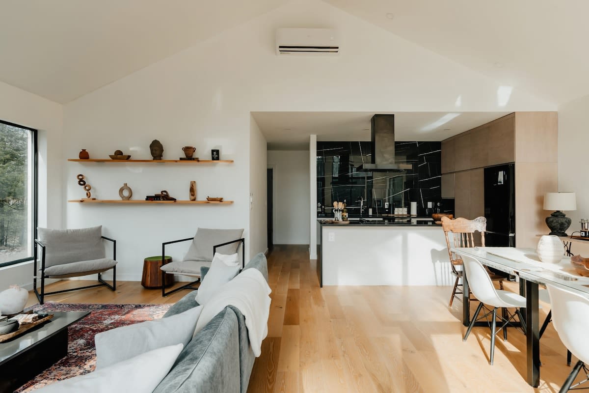 An open-concept living space with a vaulted ceiling, light hardwood floors, and white walls. The living area with a grey sofa transitions into a modern kitchen with black countertops and light wood cabinetry, and a dining area beyond.