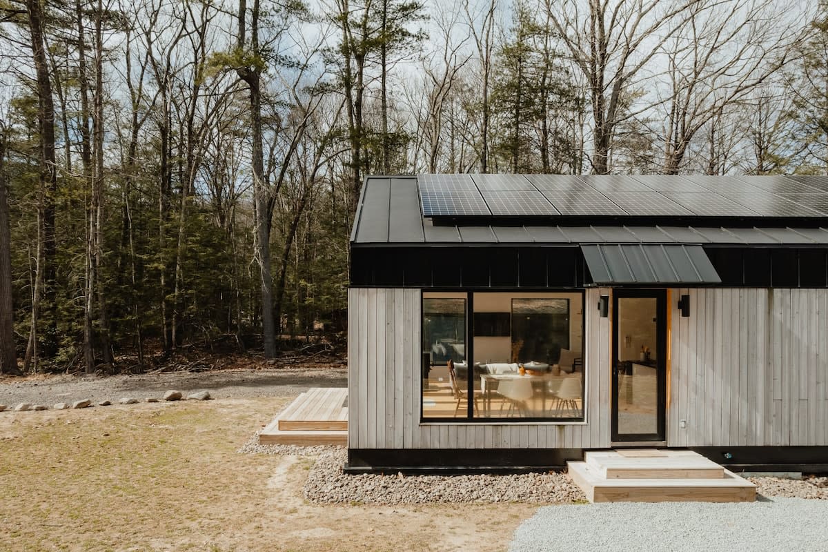 A contemporary cabin with light wood siding and a black metal roof, featuring solar panels and large windows, is nestled in a natural, wooded environment with a small wooden deck extending from the side.