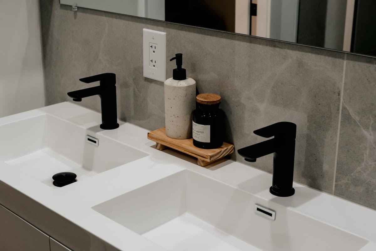 Close-up of the contemporary double vanity in the bathroom, featuring two white rectangular sinks with matte black faucets. A light gray tiled backsplash extends behind the sinks, with a wooden tray holding soap dispensers.