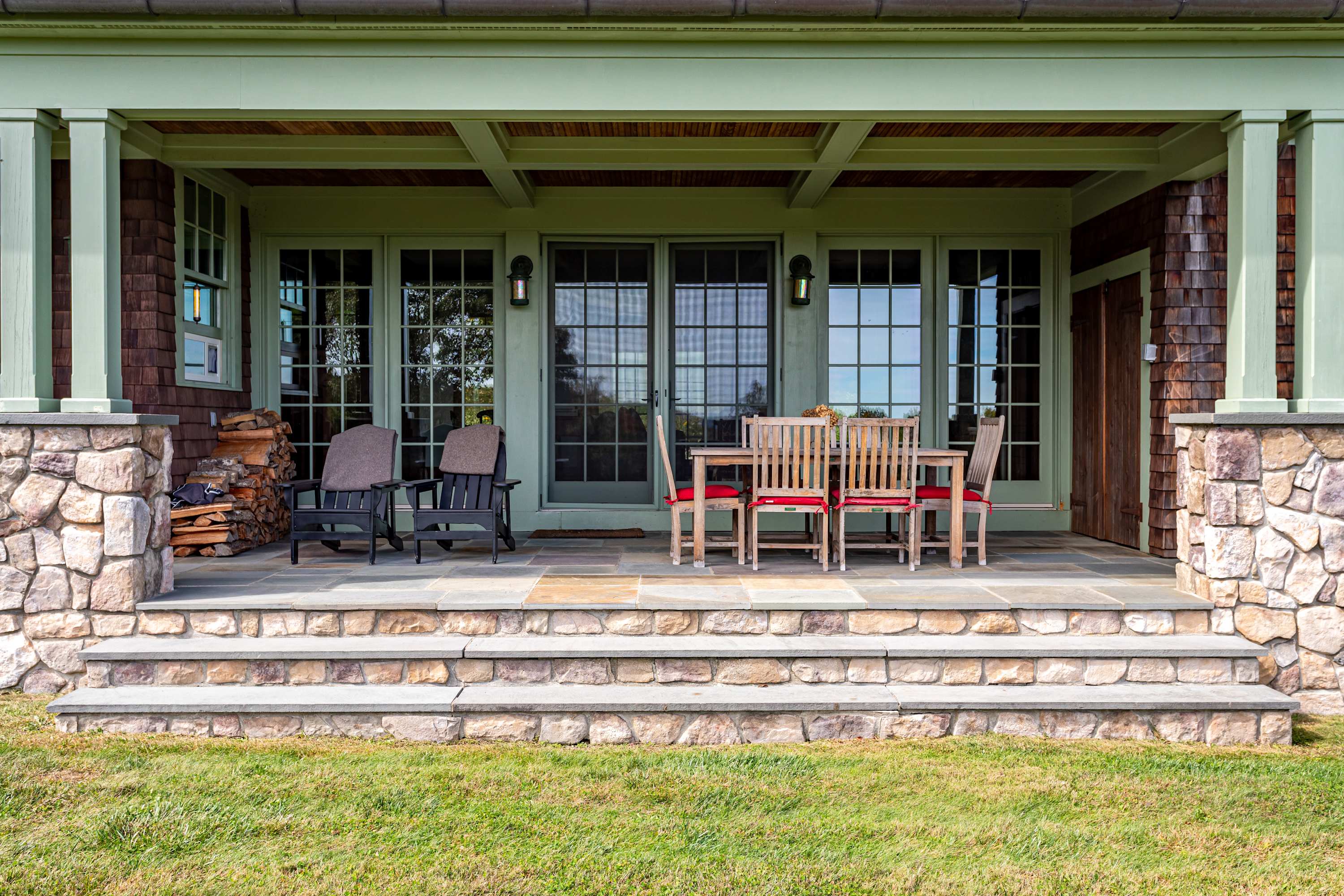 A spacious covered porch with sturdy stone pillars and a warm wooden ceiling provides an ideal outdoor living space. It features an outdoor dining set and additional seating, overlooking a manicured grassy yard with stacked firewood nearby.