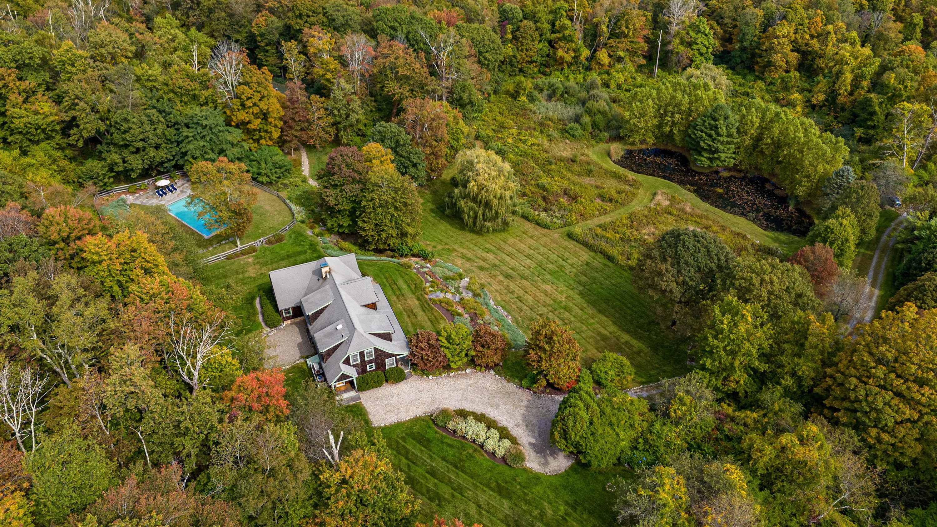 A high aerial view reveals the entire property, featuring the house, a rectangular swimming pool, a winding gravel driveway, and a pond, all set within a vast expanse of vibrant green and autumnal forest.