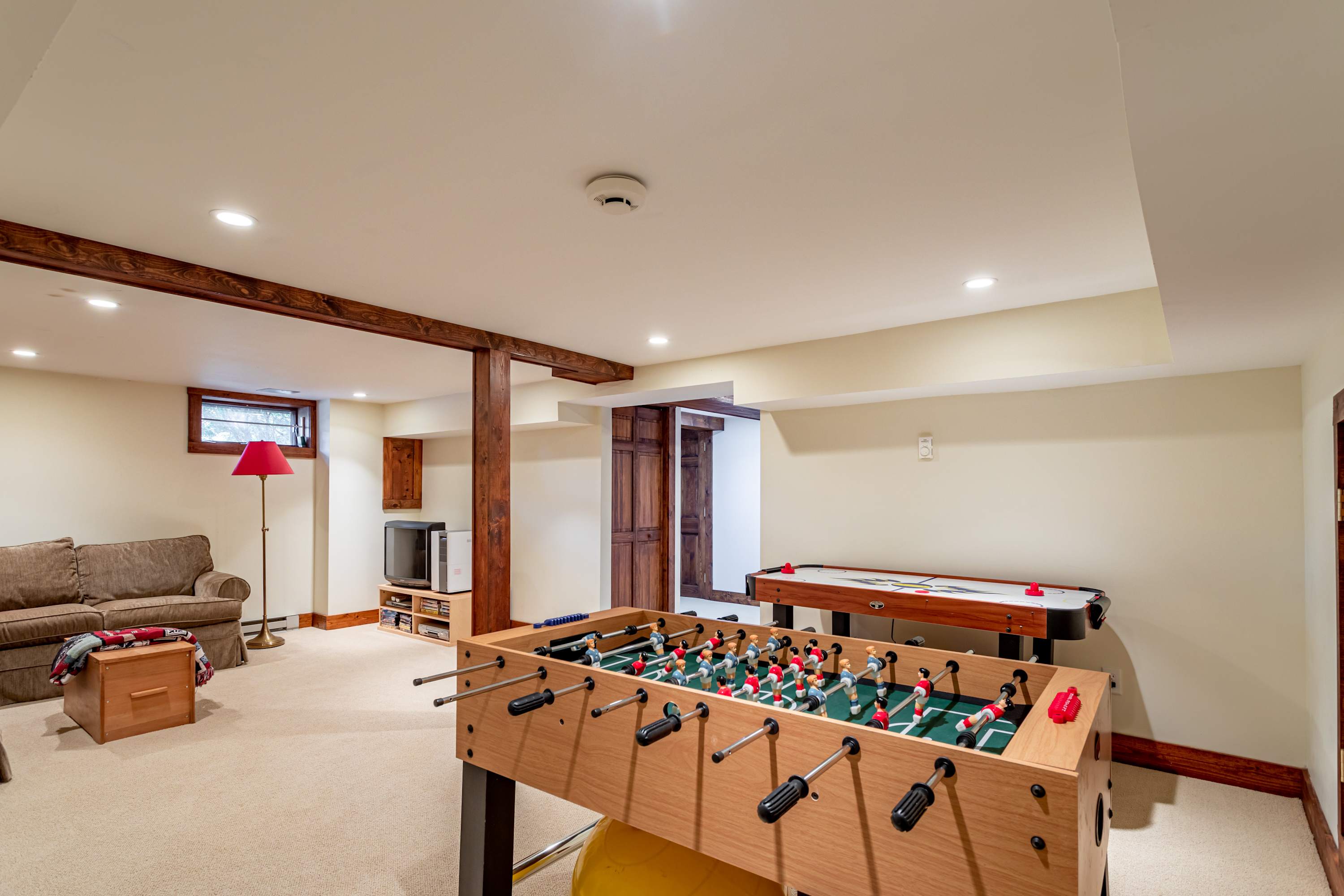 Bright recreation room featuring a foosball table and an air hockey table on a carpeted floor. A comfortable sofa and a small TV setup are visible in the background, with exposed wooden beams adding character.