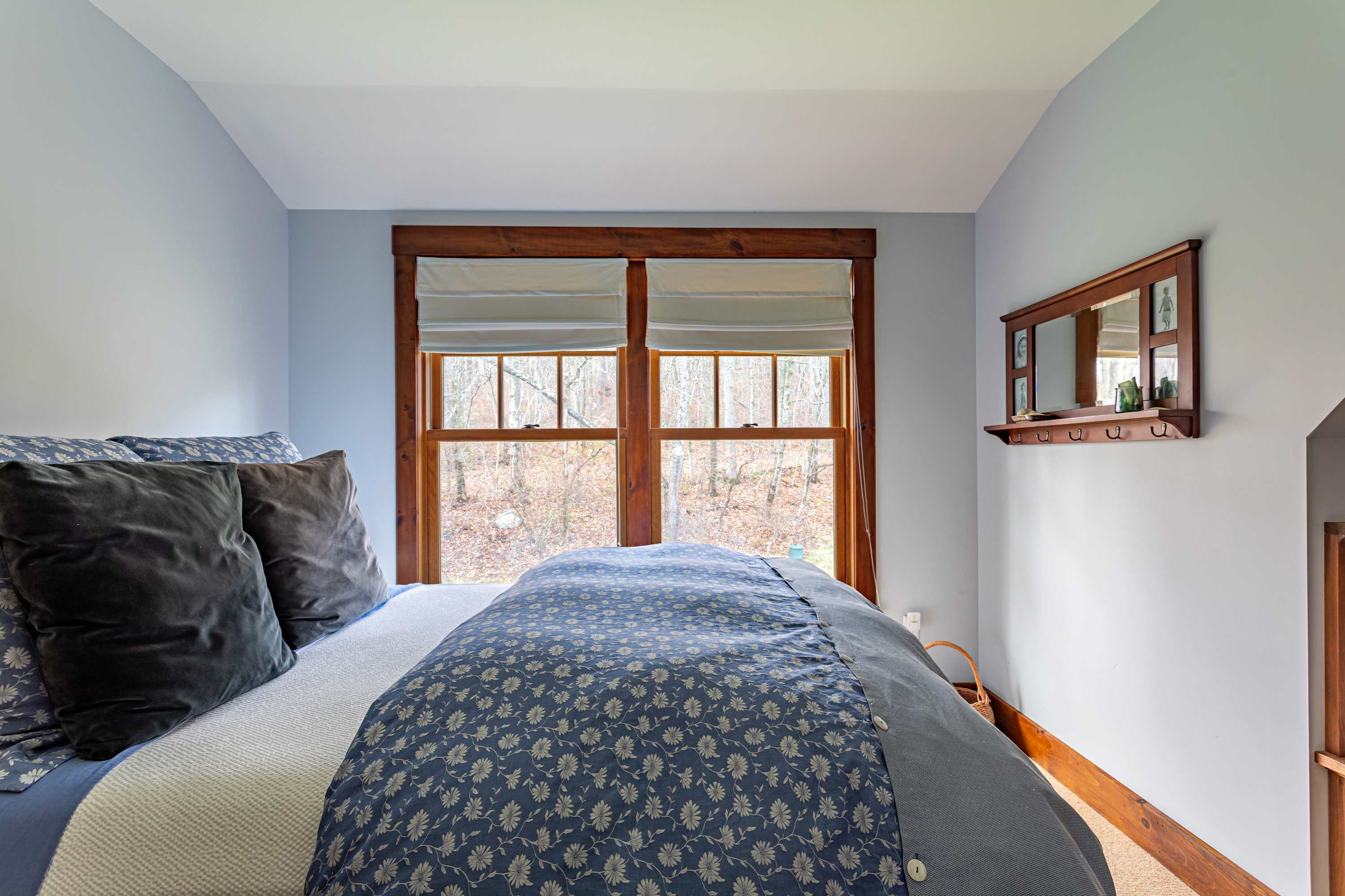 Cozy bedroom featuring a bed with blue and white floral bedding, light blue walls, and a large window with wooden frames overlooking a wooded area. A small wooden mirror and shelf are mounted on the right wall.