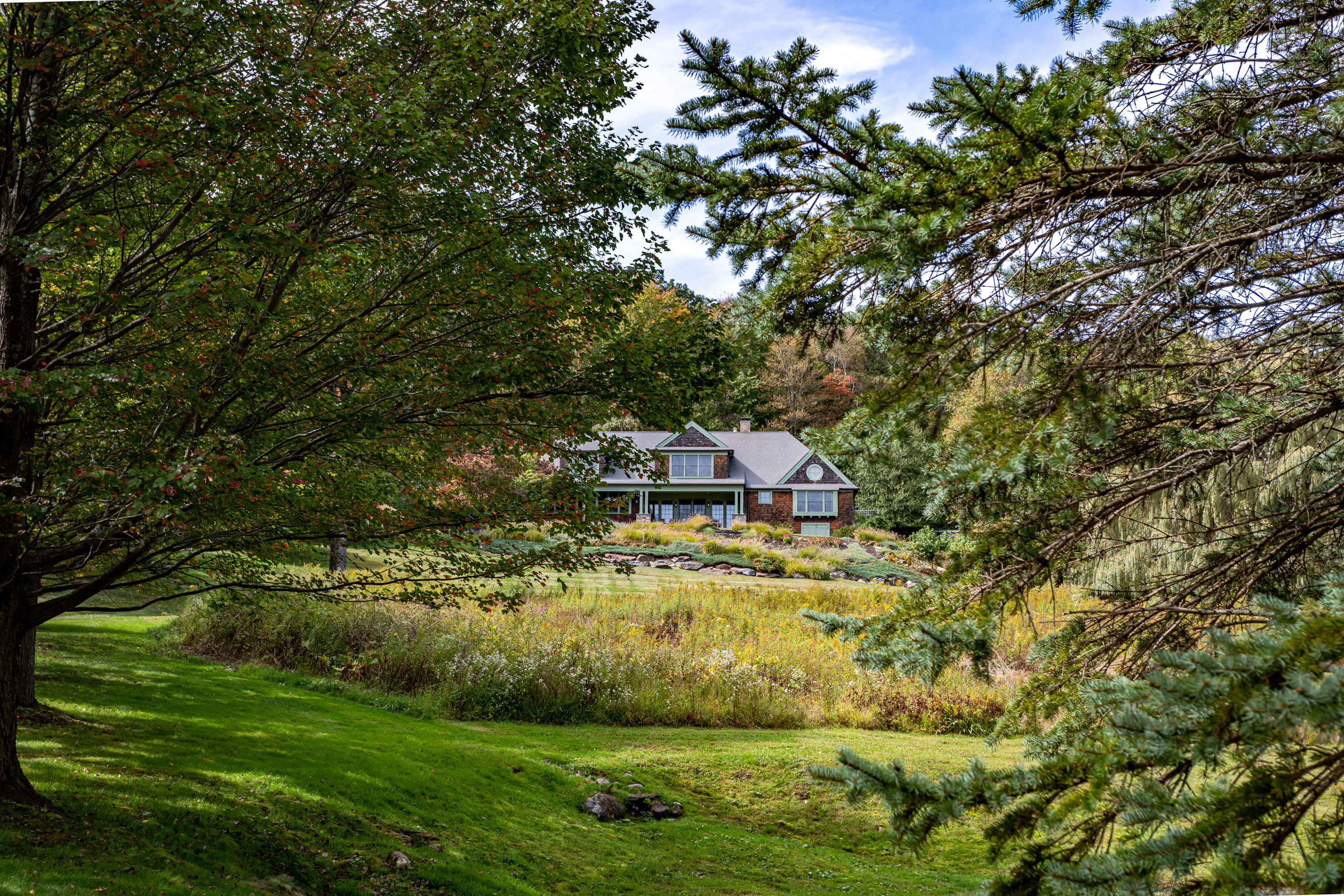 The shingle-sided house is visible in the distance through a frame of green tree branches, overlooking a wide, gently sloping grassy field with wild grasses and trees.