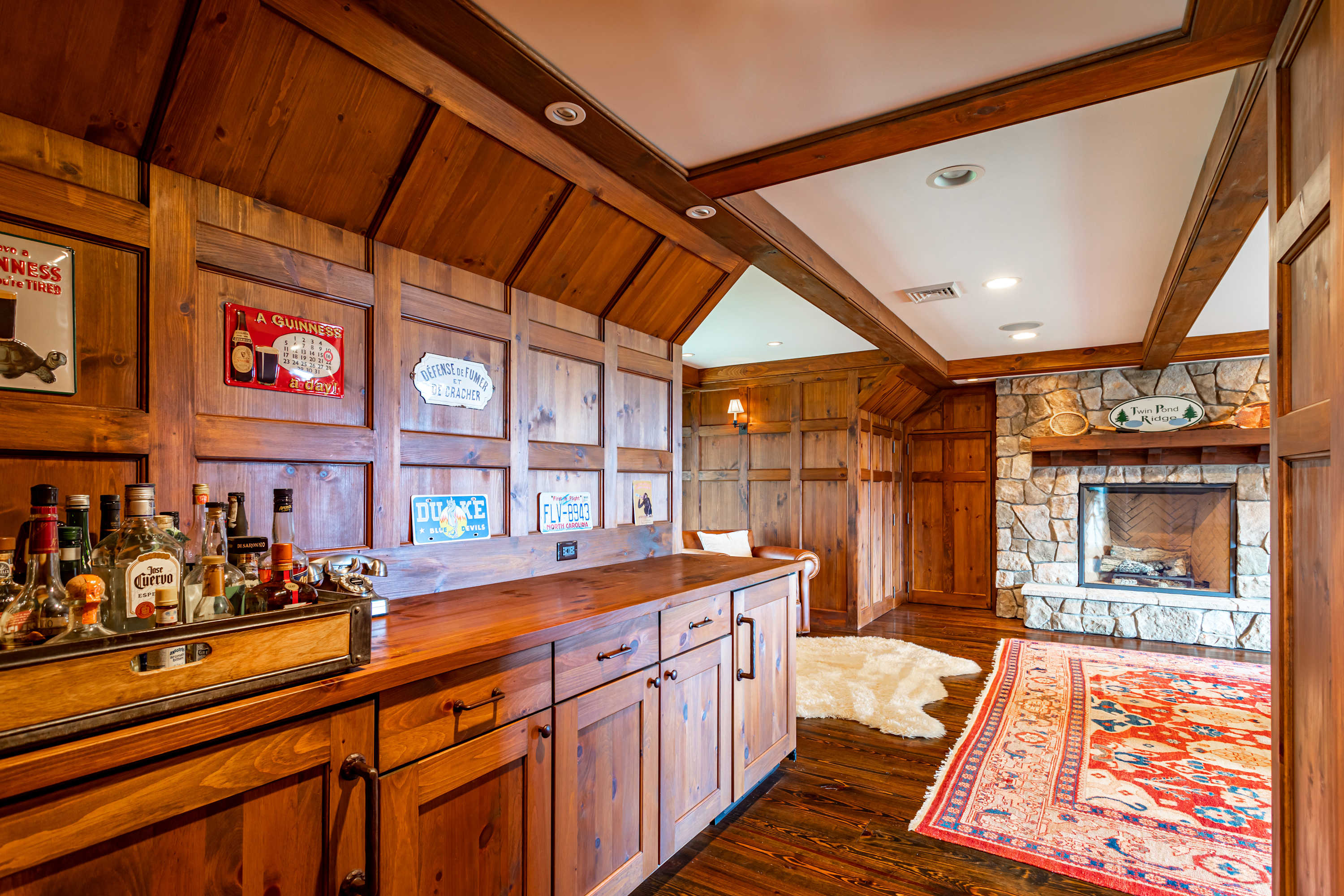 Rustic bar area with extensive wood paneling and a wooden countertop displaying various liquor bottles. A large stone fireplace with a mantel is visible on the right, adding warmth to the space.