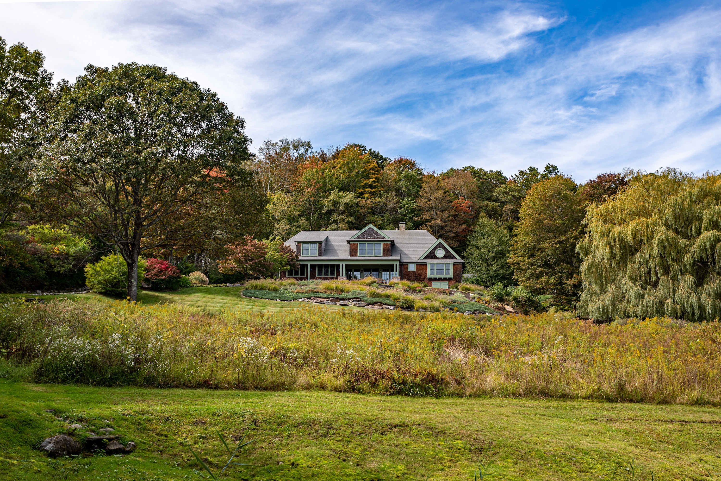A charming multi-story house with green siding and a gray roof is beautifully situated amidst a backdrop of dense, colorful autumn trees, overlooking a sprawling grassy field dotted with wildflowers under a bright blue sky.